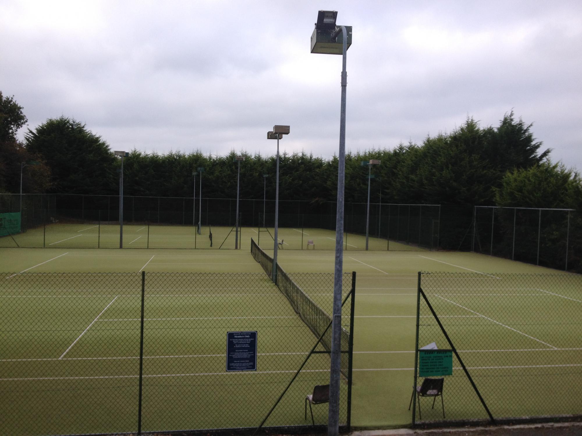 The floodlit all-weather tennis courts at Mallow Golf Club, fenced and sheltered by a line of tall evergreens